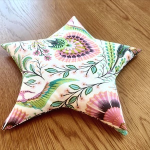 May include: A star-shaped decorative pillow with a floral pattern. The pillow features a white background with pink, green, and blue floral and leaf designs. The pillow is on a wooden surface.