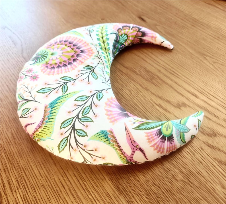 May include: A crescent-shaped pillow with a floral pattern in pink, green, and blue on a white background. The pillow is made of fabric and rests on a wooden surface. The design includes stylized flowers and leaves.
