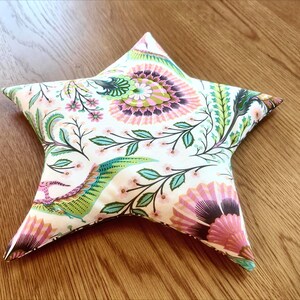 May include: A star-shaped decorative pillow with a floral pattern. The pillow features a white background with pink, green, and blue floral and leaf designs. The pillow is on a wooden surface.