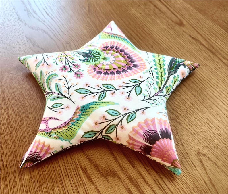 May include: A star-shaped decorative pillow with a floral pattern. The pillow features a white background with pink, green, and blue floral and leaf designs. The pillow is on a wooden surface.