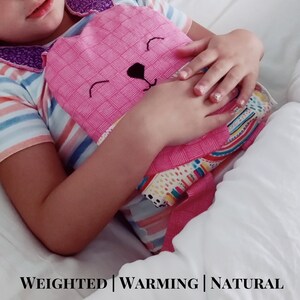 May include: A pink, heart-shaped weighted warming pad with a bear face design. The pad is held by a person wearing a striped shirt. The text "WEIGHTED | WARMING | NATURAL" is at the bottom of the image.