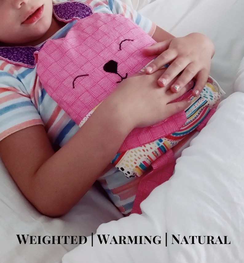 May include: A pink, heart-shaped weighted warming pad with a bear face design. The pad is held by a person wearing a striped shirt. The text "WEIGHTED | WARMING | NATURAL" is at the bottom of the image.