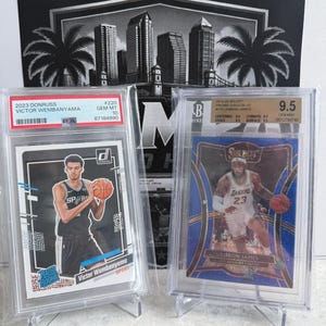May include: Two graded basketball trading cards in protective plastic cases. The left card is a 2023 Donruss Victor Wembanyama Rated Rookie PSA Gem Mt 10. The right card is a 2014-20 Select Prizm Disco Blue LeBron James BGS Gem Mint 9.5.