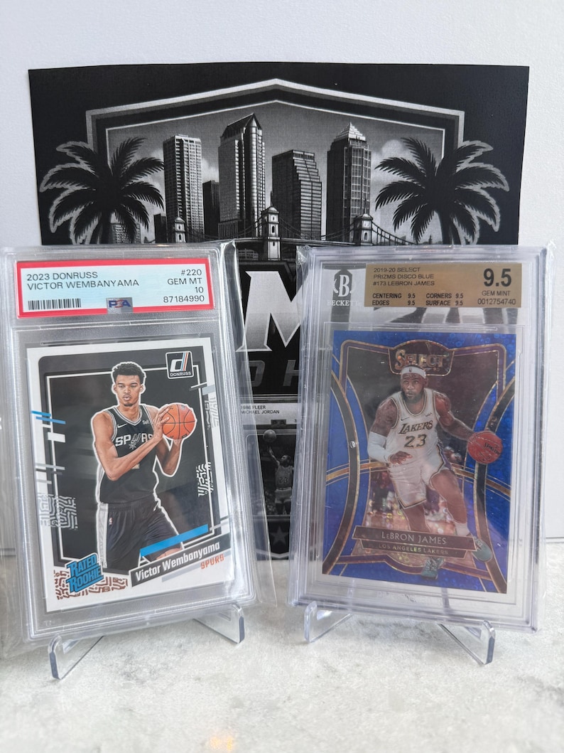 May include: Two graded basketball trading cards in protective plastic cases. The left card is a 2023 Donruss Victor Wembanyama Rated Rookie PSA Gem Mt 10. The right card is a 2014-20 Select Prizm Disco Blue LeBron James BGS Gem Mint 9.5.