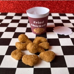 May include: A white paper cup with the words "Fried Chicken" printed on it, filled with fried chicken pieces. The cup is sitting on a black and white checkered surface. There is an empty white paper cup lid next to the full cup.