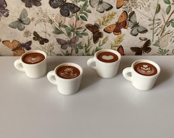 May include: Four white ceramic coffee mugs with different latte art designs. The mugs are arranged in a row on a white surface.