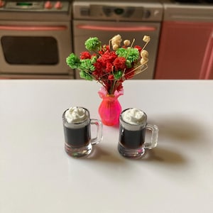 May include: Two miniature glass mugs filled with a dark liquid topped with white whipped cream. The mugs are on a white surface in front of a pink vase with a bouquet of red and green flowers.