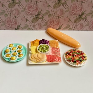 May include: Miniature food set featuring a baguette, deviled eggs on a teal plate, a charcuterie board with cheese, olives, and meats, and a small pizza. The background is a floral pattern with pink roses.