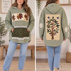 May include: A sage green quilted hooded sweatshirt featuring a front panel with three fabric autumn leaves, a dark green floral pocket with white lace trim, and a back panel displaying a large applique tree with birds and white lace accents.
