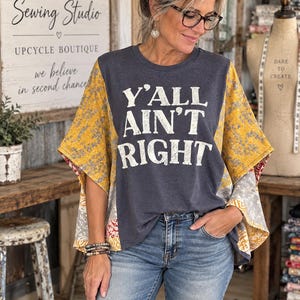May include: A charcoal gray short-sleeve top with a round neckline and wide, loose-fitting sleeves. The front features the white text Y'ALL AIN'T RIGHT. The sleeves and side panels are made of a patchwork fabric with yellow, gray, and red patterns.