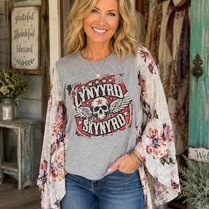 May include: A grey short-sleeved t-shirt featuring a graphic of a skull with wings and the text Lynyrd Skynyrd, paired with long, flared sleeves in a white fabric with a multi-colored floral pattern, worn with blue denim jeans.