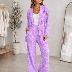 May include: A lavender two-piece set consisting of a long-sleeved open-front jacket and wide-leg trousers. Both pieces feature a textured dot pattern and fringe trim along the jacket lapels, cuffs, and the hem of the trousers.