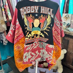 May include: A black t-shirt featuring a graphic of Bobby Hill with wings and flames, accompanied by the text "BOBBY HILL" and "FREE SPIRIT." The shirt is constructed with patchwork panels of orange, red, and white fabric featuring floral and paisley patterns.