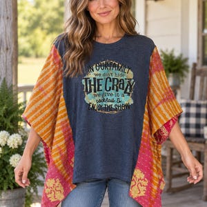 May include: A dark gray short-sleeve top with a central graphic featuring the text IN OUR FAMILY we don't hide THE CRAZY we give it a cocktail & ENJOY THE SHOW. The top has wide, loose-fitting sleeves in orange and pink with a patterned design.