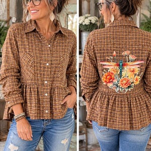 May include: A brown plaid button-down shirt with a peplum hem and long sleeves. The back features a colorful graphic print of a blue dragonfly surrounded by orange and yellow flowers. The shirt is paired with light blue distressed denim jeans.