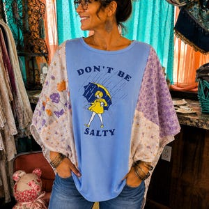 May include: A light blue short-sleeve top with a graphic print of a person holding a yellow umbrella in the rain, featuring the text "DON'T BE SALTY". The garment has wide, loose-fitting sleeves made of a patchwork fabric with floral patterns.