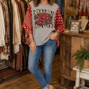 May include: A grey short-sleeved t-shirt with red patterned sleeves and a graphic print on the front featuring red roses and the text ROCK & ROLL AND ROCK. The shirt is paired with blue denim jeans and light brown ankle boots with side cutouts.