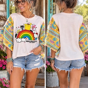 May include: A cream-colored short-sleeve t-shirt featuring a graphic of Hello Kitty and friends above a rainbow. The sleeves have a yellow, blue, and pink paisley pattern. The shirt is paired with distressed blue denim shorts with a frayed hem.