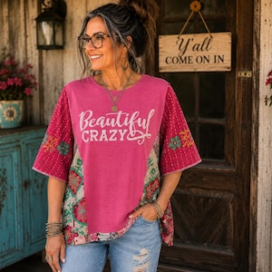 May include: A short-sleeved shirt featuring a solid pink center panel with the white text Beautiful CRAZY and side panels with a multi-colored floral pattern in shades of red, green, and cream, paired with light blue distressed denim jeans.