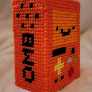May include: Orange, handmade, pixelated cube with the word "BMO" in black block letters on one side. The cube features a smiling face and other pixel art details. A unique, handcrafted item.