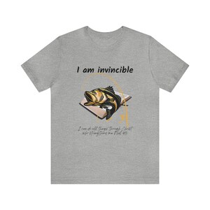 Puede incluir: Una camiseta gris con el texto "I am invincible" y un gráfico de un pez saltando de un libro con el texto "I can do all things through Christ who strengthens me Phil 4:13".