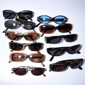 May include: A collection of sunglasses in various styles and colors, including black, brown, and gold frames. Some sunglasses have decorative embellishments. The lenses are tinted in shades of black, brown, and blue. The sunglasses are arranged on a white surface.