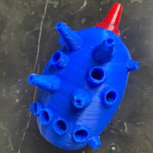 Op de afbeelding: Een blauw 3D-geprint object met een rode bovenkant en meerdere gaten. Het object is gevormd als een fles met een puntige bovenkant.