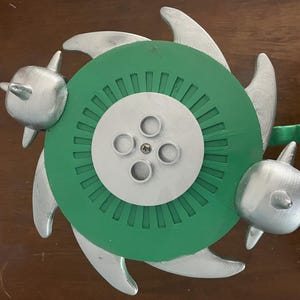 以下が含まれることがあります： 中央に緑色の円盤、銀色の円形の中央部、3つの銀色のヒレのような突起を備えた、多色の3Dプリントオブジェクト。2つの銀色のイルカ型の要素が緑色の円盤に取り付けられています。小さな緑色のハンドルも付いています。