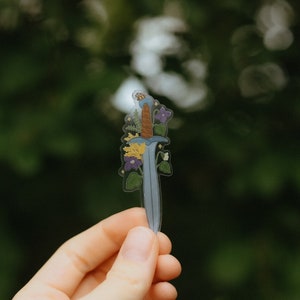 May include: A clear sticker of a blue sword with a brown handle, surrounded by yellow and purple flowers. The sticker is being held by a hand against a green background.