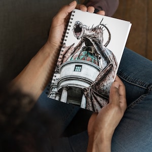 Puede incluir: Cuaderno de espiral con una ilustración detallada de un dragón posado sobre un edificio con techo abovedado. El dragón es marrón con alas abiertas y boca abierta. El edificio tiene columnas blancas y una pequeña ventana.