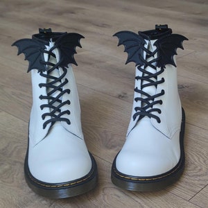 Peut inclure: Bottes blanches en cuir à lacets avec des breloques en forme de chauve-souris noires. Les bottes ont une semelle noire et des coutures jaunes.