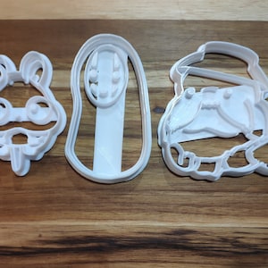 May include: Three white plastic cookie cutters in various shapes. The shapes include a frog, a long oval with circular details, and a character wearing a hat. The cutters are on a wooden surface.