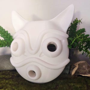 May include: A white, stylized mask with large, circular eye and mouth openings, and pointed ears. The mask has a smooth surface and is likely made of a solid material. The mask is a decorative item.