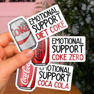 May include: Three die-cut stickers featuring illustrations of soda cans. One shows a silver Diet Coke can with the text EMOTIONAL SUPPORT DIET COKE. Another shows a red Coke Zero can with the text EMOTIONAL SUPPORT COKE ZERO. The third shows a red Coca-Cola can with the text EMOTIONAL SUPPORT COCA COLA.
