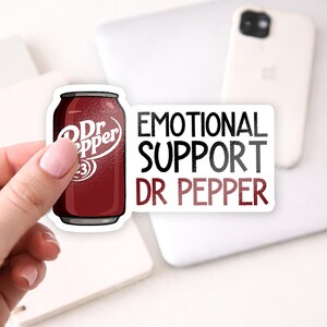 May include: A sticker featuring a Dr Pepper can and the text "EMOTIONAL SUPPORT DR PEPPER." The Dr Pepper can is a dark red color with white text. The text is in black and red.
