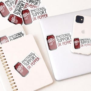 May include: A collection of stickers featuring a Dr. Pepper can design with the text "EMOTIONAL SUPPORT DR PEPPER." The stickers are placed on a white laptop, a smartphone, and a spiral notebook. The stickers are red and white.