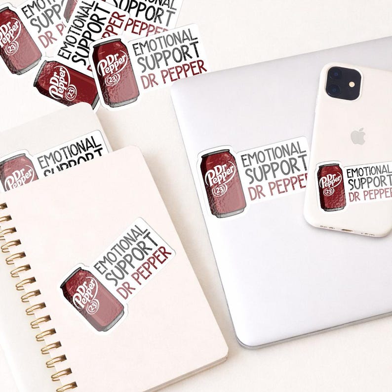 May include: A collection of stickers featuring a Dr. Pepper can design with the text "EMOTIONAL SUPPORT DR PEPPER." The stickers are placed on a white laptop, a smartphone, and a spiral notebook. The stickers are red and white.