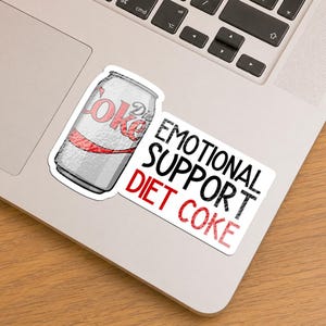 May include: A sticker featuring a silver Diet Coke soda can illustration next to the black and red text that reads EMOTIONAL SUPPORT DIET COKE. The design is printed on a white background with a die-cut border, showcasing a metallic texture.