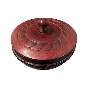 Puede incluir: Una caja redonda de madera de color rojo oscuro con una tapa a juego. La tapa presenta un pomo esférico central y un patrón decorativo tallado de líneas diagonales que irradian hacia afuera. La base muestra un patrón diagonal repetido.