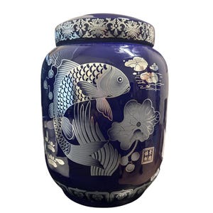 Puede incluir: Un jarrón de cerámica azul cobalto con tapa, con un diseño de pez koi y flor de loto plateado. El jarrón tiene una banda decorativa plateada alrededor de la tapa y la base. El diseño es de estilo asiático tradicional.
