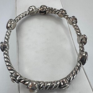 May include: Silver bangle bracelet with a twisted rope design. The bracelet has several decorative elements, including small, round embellishments and a single pearl. The bracelet is shown on a white surface.