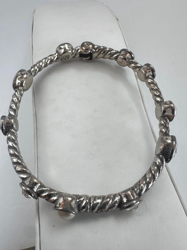 May include: Silver bangle bracelet with a twisted rope design. The bracelet has several decorative elements, including small, round embellishments and a single pearl. The bracelet is shown on a white surface.
