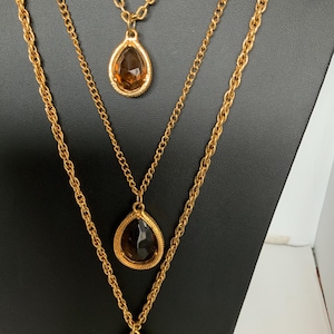 May include: Three gold-toned necklaces with teardrop-shaped pendants. Each pendant has a brown, faceted stone. The chains are of varying lengths and styles, displayed on a black stand.