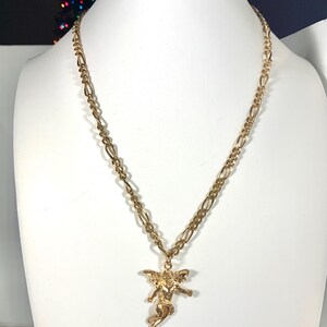 May include: A gold-colored necklace with a pendant of an angel. The necklace has a figaro chain and is displayed on a white mannequin. The angel pendant is detailed with wings and outstretched arms.