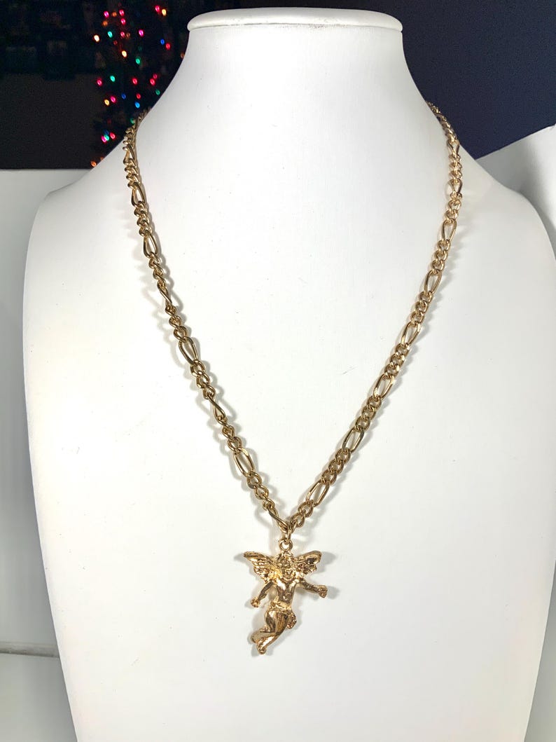 May include: A gold-colored necklace with a pendant of an angel. The necklace has a figaro chain and is displayed on a white mannequin. The angel pendant is detailed with wings and outstretched arms.