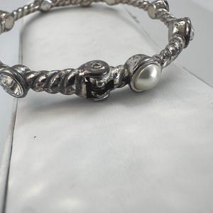 May include: Silver-coloured bangle bracelet with a twisted rope design. The bracelet features clear crystals and a faux pearl. The bracelet is a piece of jewellery.