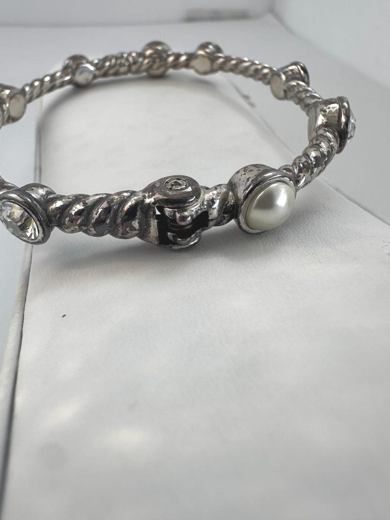 May include: Silver-coloured bangle bracelet with a twisted rope design. The bracelet features clear crystals and a faux pearl. The bracelet is a piece of jewellery.