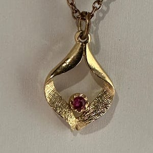 May include: Gold-toned pendant necklace with a small, round, red gemstone at the center. The pendant has a textured, openwork design. The chain is a delicate link style.