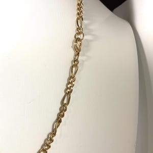 May include: A gold-colored chain necklace with a Figaro link design. The necklace is displayed on a white mannequin. The chain has a repeating pattern of three small circular links followed by one elongated oval link.