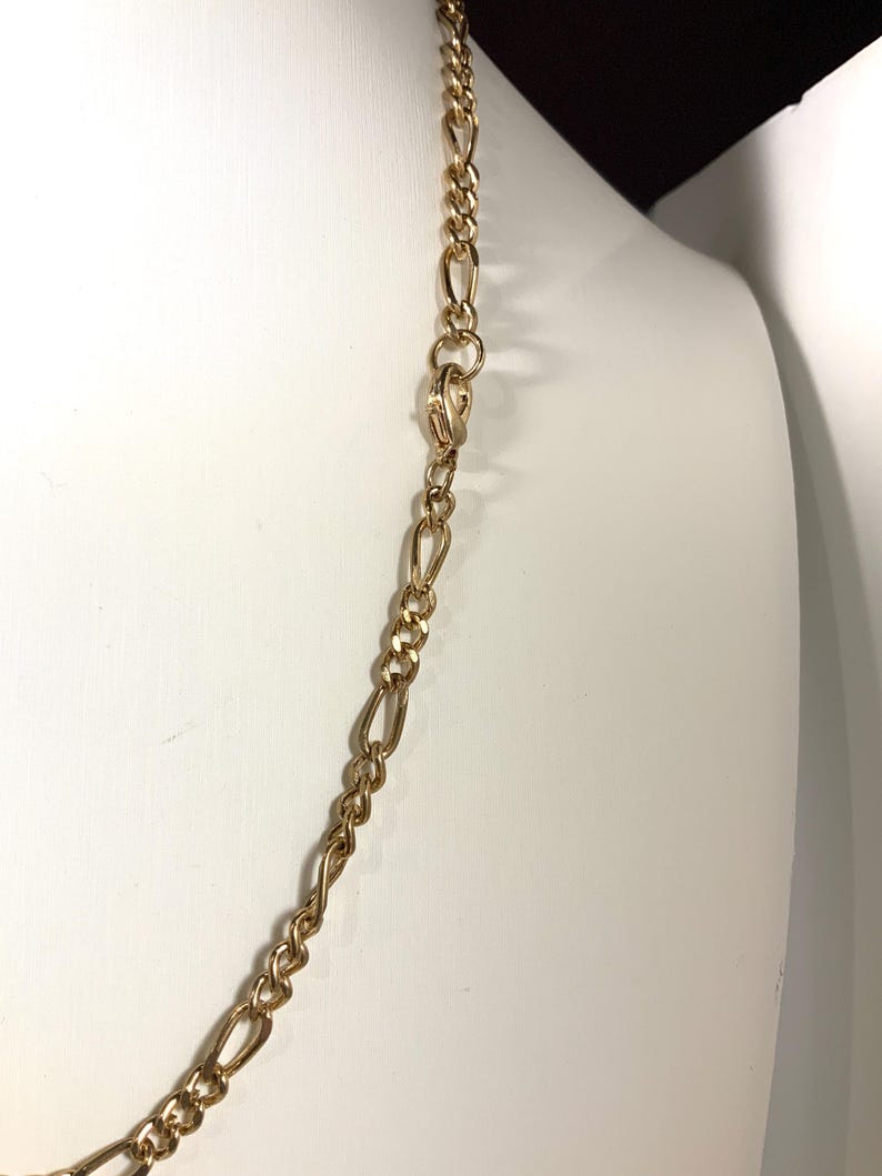 May include: A gold-colored chain necklace with a Figaro link design. The necklace is displayed on a white mannequin. The chain has a repeating pattern of three small circular links followed by one elongated oval link.
